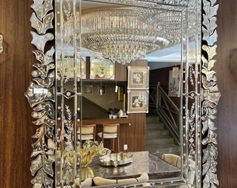 May include: Rectangular mirror with an ornate, etched glass frame. The frame features a leaf and floral design. The mirror reflects a room with a chandelier, dining table, and staircase. The mirror is mounted on a dark wood wall.