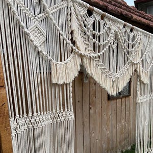 May include: Cream-coloured macrame wall hanging with intricate geometric and fringe details. The macrame is suspended from a wooden rod, creating a decorative curtain effect. The wall hanging is displayed against a wooden wall.