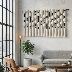 May include: A decorative wall hanging featuring white spherical shapes and tassels, interspersed with black and beige details. The artwork is suspended from a dark rod, adding a stylish touch to a contemporary living room setting.
