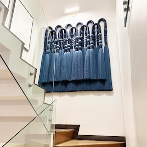 May include: A large, blue textile wall hanging with a repeating pattern of looped shapes and long tassels. The artwork is mounted on a white wall, above a wooden staircase with glass railings.