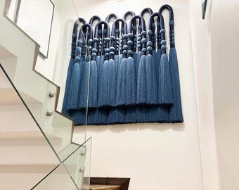 May include: A large, blue textile wall hanging with a repeating pattern of looped shapes and long tassels. The artwork is mounted on a white wall, above a wooden staircase with glass railings.