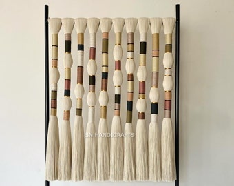 May include: A wall hanging made of woven yarn with tassels. The yarn is in shades of beige, brown, pink, and black. The hanging is mounted on a black metal frame. The text "SN HANDICRAFTS" is visible in the bottom center of the image.
