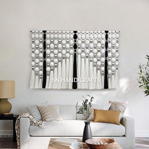 May include: A black and white macrame wall hanging with white pom poms and black tassels. The wall hanging is made of yarn and is hanging on a white wall in a living room setting.
