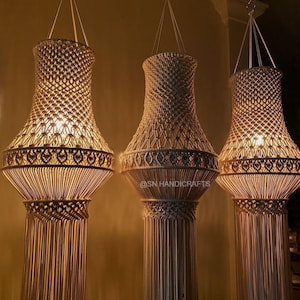 May include: Three hanging macrame lamps, each with a warm, inviting glow. The lamps feature a woven design with a flared top and a long, fringed bottom. They are suspended from the ceiling by thin cords. The lamps are a neutral colour, with a textured appearance.