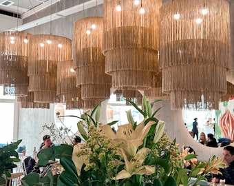 May include: A restaurant interior featuring several large, tiered, beige rope pendant lights hanging from the ceiling. A floral arrangement with white lilies and green foliage is in the foreground. People are seated at tables.