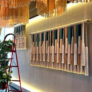 May include: A decorative wall hanging featuring vertical strands in beige, teal, and brown, creating a textured pattern. The piece is mounted on a wooden rod. Similar strands hang from the ceiling, and a red ladder with plants is in the foreground.