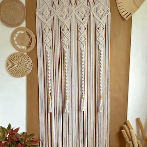 Op de afbeelding: Een witte macramé wandhanger met een geometrisch patroon en houten kralen. De hanger is gemaakt van katoenen koord en heeft een franje aan de onderkant.