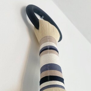Puede incluir: Un tapiz de pared hecho de fibras naturales con un anillo negro en la parte superior. El tapiz est&aacute; rayado con hilo beige, blanco, gris y negro. El texto "SN HANDICRAFTS" es visible en el tapiz.