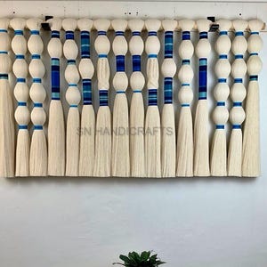 May include: A wall hanging made of white yarn with blue and turquoise accents. The yarn is tied into knots and then fanned out to create a fringe effect. The wall hanging is approximately 20 inches wide and 15 inches tall.