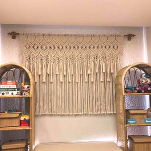 May include: A macrame wall hanging with a geometric pattern and fringe. The hanging is made of natural cotton cord and is a light beige color. It is hung on a wooden dowel rod.