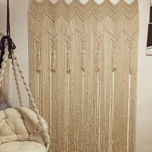 May include: A beige macrame wall hanging with a geometric design. The wall hanging is made of knotted cotton rope and hangs from a wooden rod. A hanging chair is visible in the foreground.