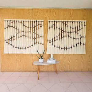 Könnte beinhalten: Zwei große, cremefarbene Wandbehänge mit einem geometrischen Design aus vertikalen Elementen und dunkelbraunen Akzenten. Die Wandbehänge sind an einer hellen Holzvertäfelung angebracht. Darunter steht ein kleiner, ovaler Tisch mit einer Pflanze und Büchern.