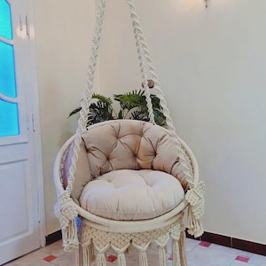 Op de afbeelding: Een beige macramé hangstoel met een rond, gecapitonneerd kussen en een bijpassend zitkussen. De stoel hangt aan een gevlochten touw en heeft decoratieve franjes. Ontworpen voor gebruik binnenshuis.