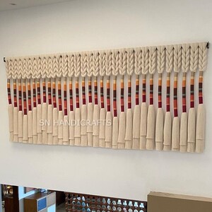 May include: A white macrame wall hanging with a braided top and long tassels. The tassels are accented with red, orange, and brown stripes. The wall hanging is labeled "SN HANDICRAFTS".
