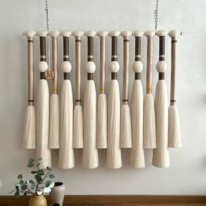 May include: A decorative wall hanging featuring multiple vertical elements with cream-colored tassels. The design includes brown and beige wrapped sections, with a wooden disc bearing the letter "M". The piece is suspended from a chain.
