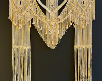May include: Cream-colored macrame wall hanging with intricate geometric patterns and fringe. The macrame is suspended from a wooden rod, featuring diamond and triangle designs. Decorative tassels hang from the bottom of the piece, adding texture and visual interest.