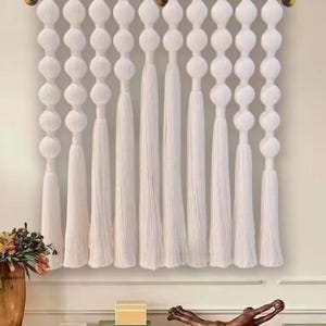 May include: White macrame wall hanging with a series of large, round knots and long tassels. The wall hanging is suspended from a wooden rod with gold-colored hardware. The design is a modern, bohemian style.