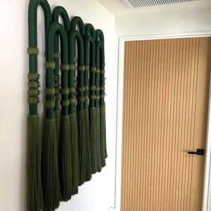 Puede incluir: Una decoración mural con múltiples elementos verdes arqueados con detalles decorativos y borlas. La obra de arte está montada en una pared blanca, con una puerta de madera con ranuras verticales en el fondo.