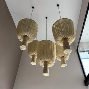 Puede incluir: Un grupo de cinco lámparas colgantes con una textura natural y tejida. Cada lámpara tiene forma de hongo, con una pantalla ancha y deshilachada y un tallo cilíndrico. Las lámparas cuelgan de cables negros.