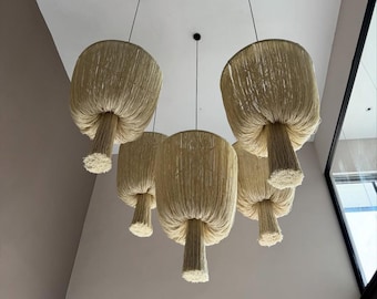 May include: A cluster of five pendant lights with a natural, woven texture. Each light fixture has a mushroom-like shape, with a wide, fringed shade and a cylindrical stem. The lights hang from black cords against a white ceiling.
