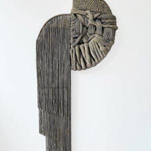 May include: A large, abstract wall hanging in shades of gray. The piece features a woven half-circle design on the right, with a knotted texture. The left side has a semi-circular shape with long, hanging fringe. A modern, textured art piece.