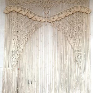 May include: A large, cream-colored macrame wall hanging. The intricate design features knotted patterns, fringe, and a scalloped valance. The piece is made of natural fibers and hangs against a white wall, adding a bohemian touch to any space.