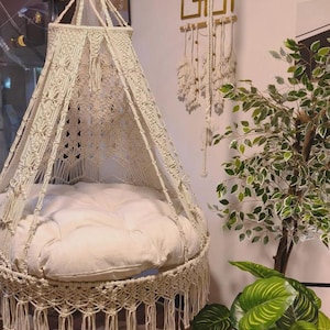 May include: Cream-colored macrame hanging chair with a round, cushioned seat. The chair features intricate woven patterns and a fringed bottom edge. The chair is suspended from a ceiling hook, creating a cozy, bohemian-style seating area.