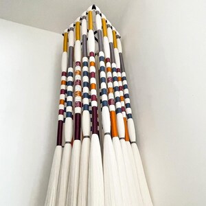 May include: A decorative wall hanging composed of multiple broom-like elements. Each element features a white brush head and a handle with alternating stripes of color, including orange, blue, and burgundy. The handles have the text "SN HANDICRAFTS".