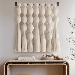 Könnte beinhalten: Ein weißer Makramee-Wandbehang mit einer Holzleiste oben. Der Wandbehang besteht aus einer Reihe von geknüpften Schnüren, die ein Muster aus Kreisen bilden. Der Wandbehang hängt an einer weißen Wand.
