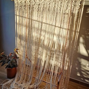 Puede incluir: Un tapiz de macramé en algodón color crema. El intrincado diseño presenta un patrón geométrico en la parte superior y hebras largas y fluidas. La pieza se exhibe en un marco de metal color cobre, creando una estética bohemia. Una planta en maceta es visible a la izquierda.