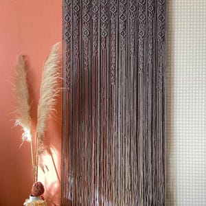 May include: A macrame curtain in shades of gray hangs against a peach-colored wall and a patterned wallpaper. The curtain features intricate knotted patterns at the top and long, vertical strands. Decorative pampas grass and a woven vase are in the foreground.