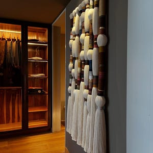 May include: A large, vertical wall hanging with a series of cylindrical elements and white tassels. The cylinders are wrapped in various colors and textures, creating a layered effect. The piece is mounted on a gray wall, adding a touch of texture and visual interest to the space.