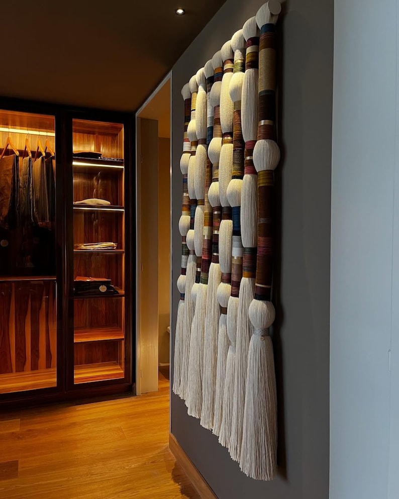 May include: A large, vertical wall hanging with a series of cylindrical elements and white tassels. The cylinders are wrapped in various colors and textures, creating a layered effect. The piece is mounted on a gray wall, adding a touch of texture and visual interest to the space.