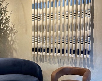 May include: A white, blue, and black wall hanging made of yarn with a fringe at the bottom. The wall hanging is made of multiple strands of yarn that are woven together to create a pattern. The wall hanging is hanging on a wall in a room with a blue couch and a brown leather chair.