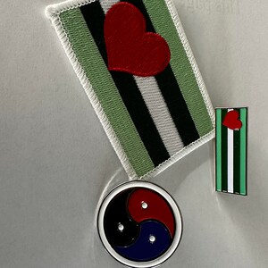 Könnte beinhalten: Drei gestickte Patches mit verschiedenen Designs. Der erste Patch ist grün, weiß und schwarz mit einem roten Herzen in der Mitte. Der zweite Patch ist grün, weiß und schwarz mit einem roten Herzen in der Mitte. Der dritte Patch ist ein schwarzer, roter und blauer Kreis mit einem weißen Umriss.