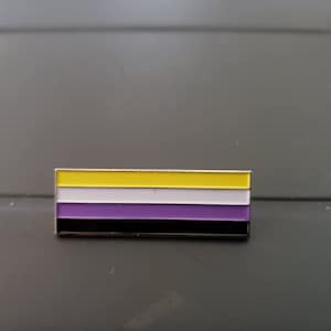 Puede incluir: Un pin de metal con un diseño de rayas amarillas, blancas, moradas y negras. El pin es una representación de la bandera del orgullo no binario.