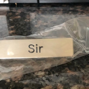 Könnte beinhalten: Ein rechteckiges Namensschild aus gebürstetem Metall mit dem eingravierten Wort "Sir" in einer dunklen, serifenlosen Schrift. Das Namensschild befindet sich in einer durchsichtigen Kunststoffhülle, die ein sauberes und funktionales Erscheinungsbild bewahrt.