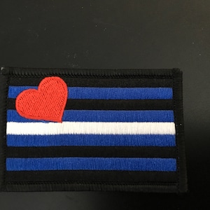 May include: A black and blue embroidered patch with a red heart in the top left corner. The patch has a white stripe in the middle and is surrounded by a black border.