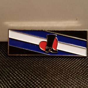May include: A silver pin with a blue, white, and black striped flag design. A red heart with a black boot is in the center of the flag.