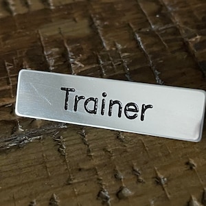 Pode incluir: Uma etiqueta de nome de metal prateado com a palavra "Trainer" impressa em letras pretas.