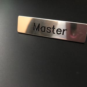Puede incluir: Una placa rectangular de metal cepillado con la palabra "Master" grabada en una fuente sans-serif. La placa de identificación está colocada sobre un fondo oscuro, posiblemente negro.