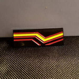 May include: A black enamel pin with a red and yellow chevron design.