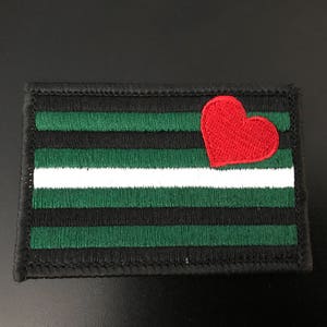 May include: Embroidered patch with a design featuring horizontal stripes in black, green, and white, with a red heart in the upper right corner. The patch has a black border and is likely for clothing or accessories.
