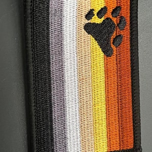 May include: A black, brown, tan, white, and gray pride flag with a black bear paw print in the center.