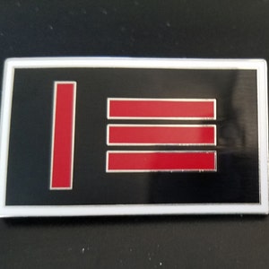 Puede incluir: Un pin de esmalte negro y rojo con borde blanco. El pin presenta tres barras rojas horizontales y una barra roja vertical.