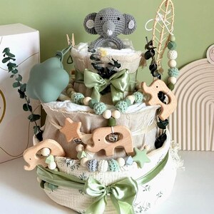 May include: A three-tiered baby diaper cake decorated with green bows, wooden elephant and star accents, and a crocheted gray elephant topper. The cake is adorned with a garland of wooden beads and green accents.