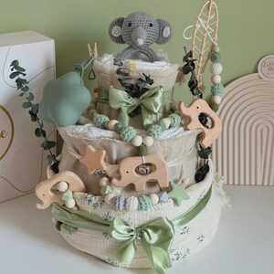 May include: A three-tiered baby diaper cake with a gray crocheted elephant topper. The cake is decorated with green ribbons, wooden elephant and star accents, and a beaded garland. A box with the text "Lulis Made" is visible in the background.