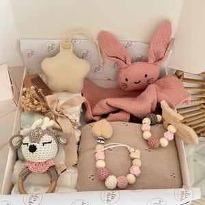 May include: A baby gift set in a white box, featuring a pink bunny plush, a star-shaped teether, a wooden deer rattle, a pacifier clip, and a blanket. The set includes a wooden comb and other baby essentials. The items are in shades of pink, beige, and white.