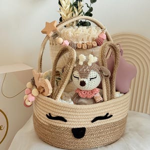 Könnte beinhalten: Ein beigefarbener und brauner gewebter Korb mit einem lächelnden Gesichtsdesign, gefüllt mit Babyzubehör. Enthält ein gehäkeltes Reh, ein sternförmiges Holzspielzeug und ein sternförmiges rosa Kissen. Der Korb enthält auch dekoratives Grün.