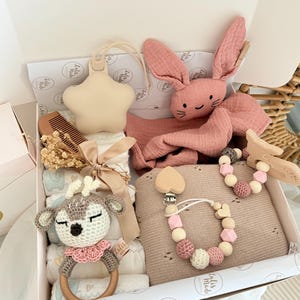 May include: A baby gift box filled with a variety of items. Includes a beige star-shaped teether, a pink plush bunny, a crochet deer rattle, a wooden comb, a pacifier clip, and a knitted blanket. The box is white with a pink interior.